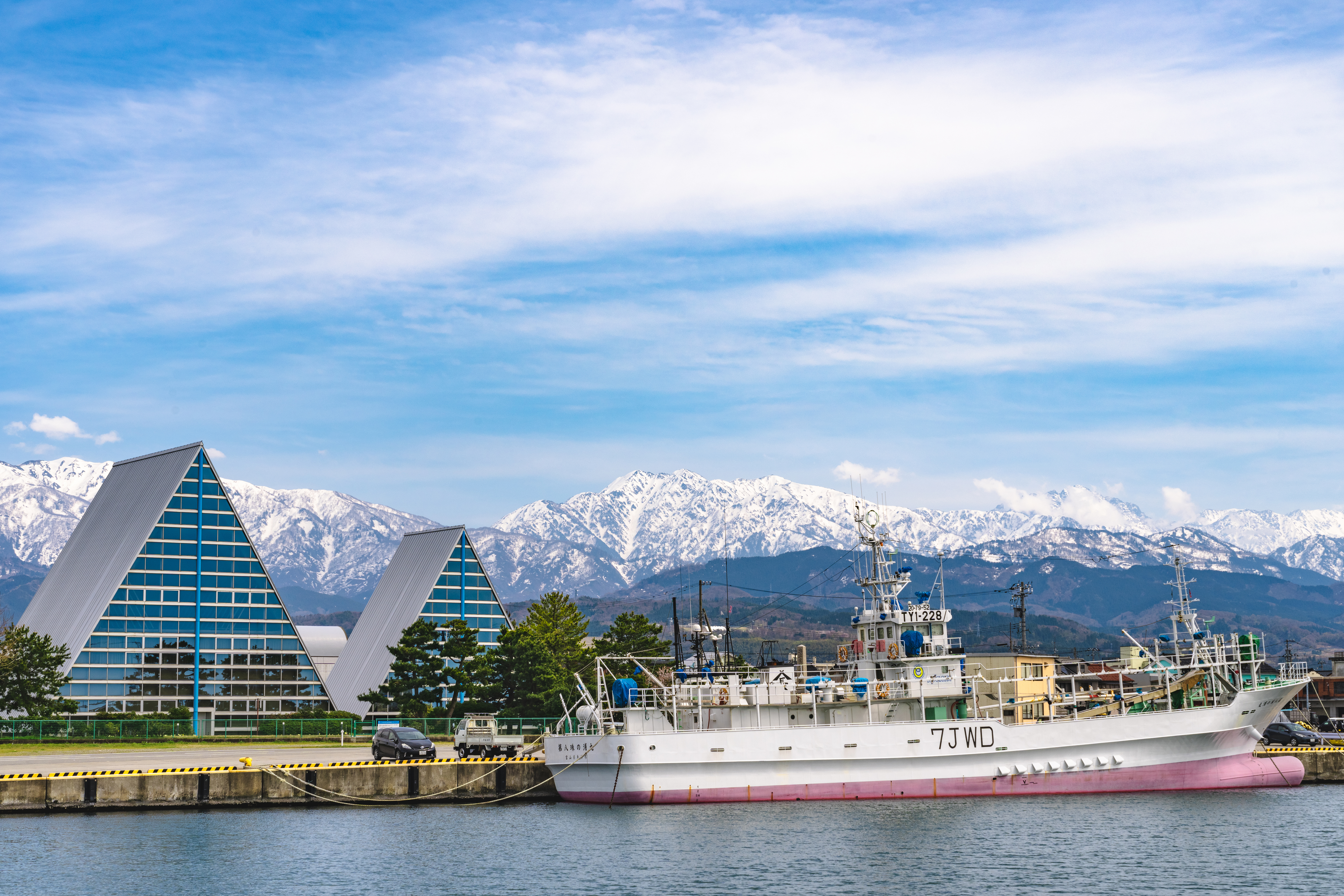 富山県魚津市の魚津漁港と立山連峰。三角形の特徴的な建物（魚津埋没林博物館）と雪をかぶった山々を背景に漁船が停泊している